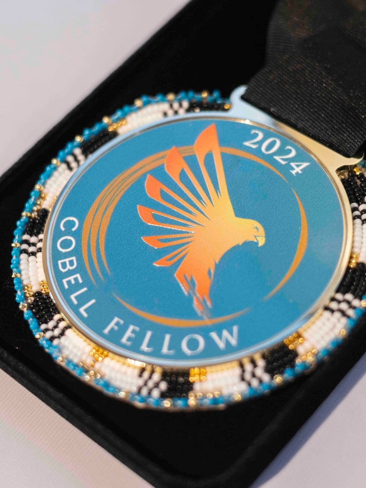The Cobell Fellow 2024 medallion — a golden eagle in flight on a blue enamel face ringed by a beaded border, resting in its presentation case
