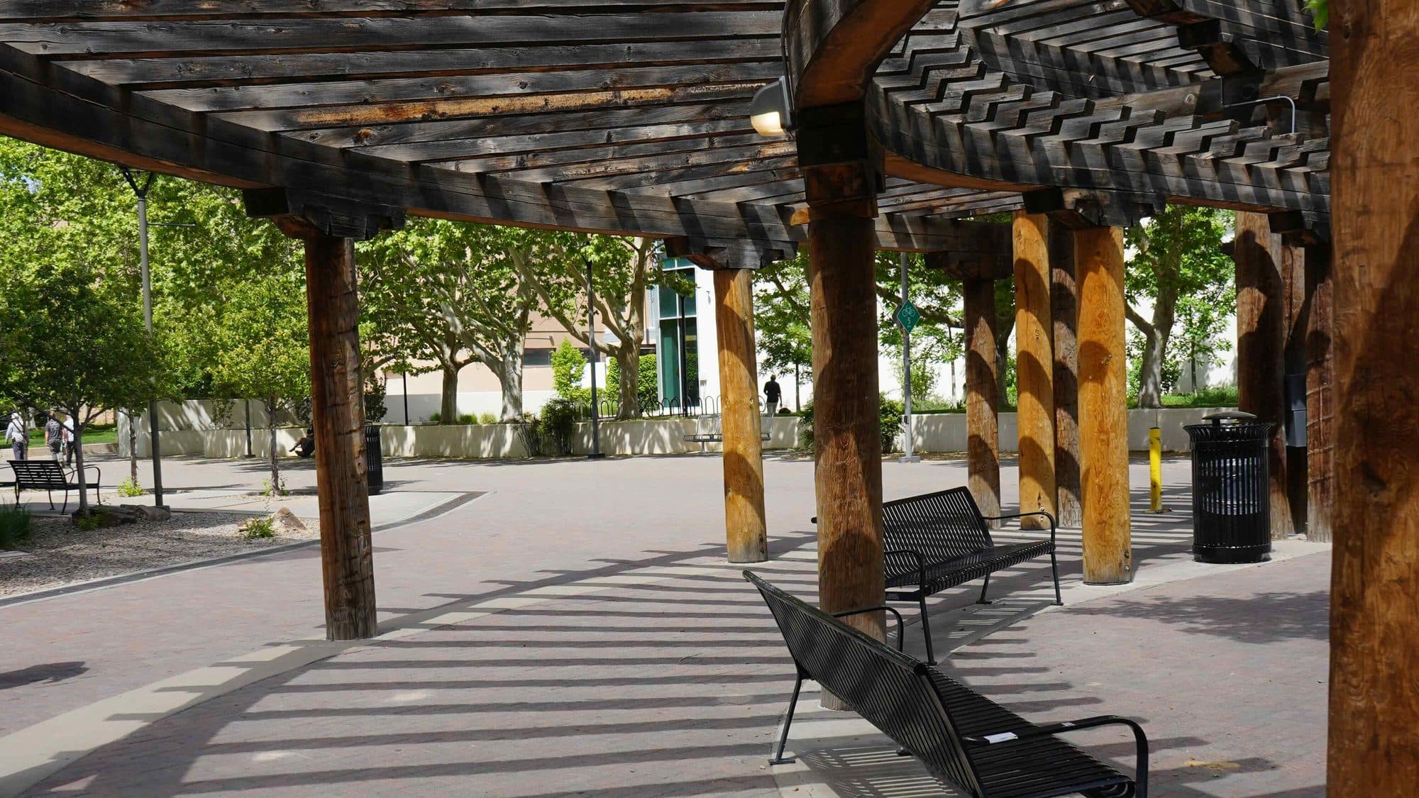 A wooden pergola casts striped shadows over a brick campus courtyard lined with benches