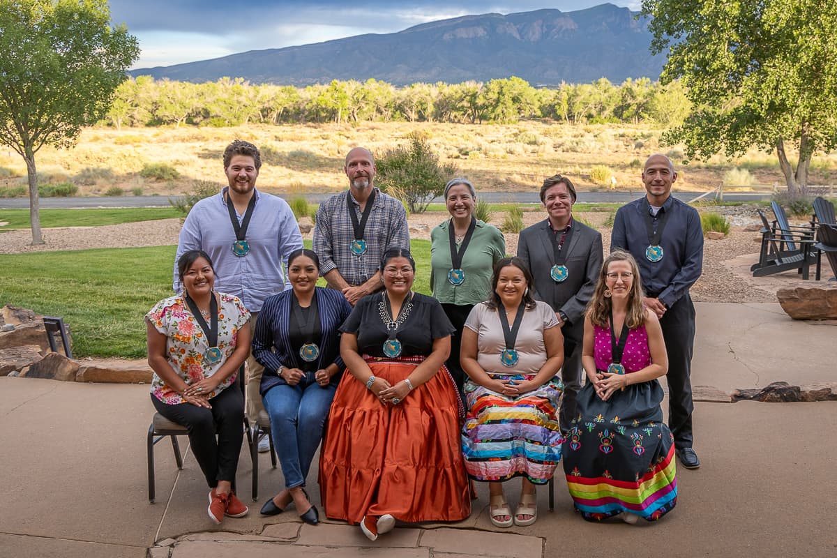 A group of Cobell Scholarship recipients wearing medallions at an outdoor gathering, with mountains behind them